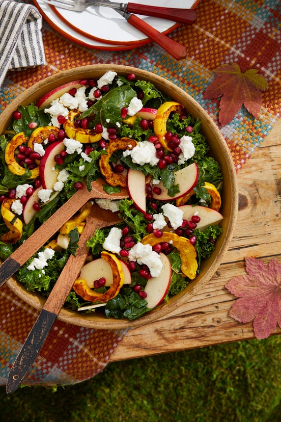 a kale delicata squash pomegranate seed apple and goat cheese salad in a wood salad bowl set on a table with a plaid tablecloth