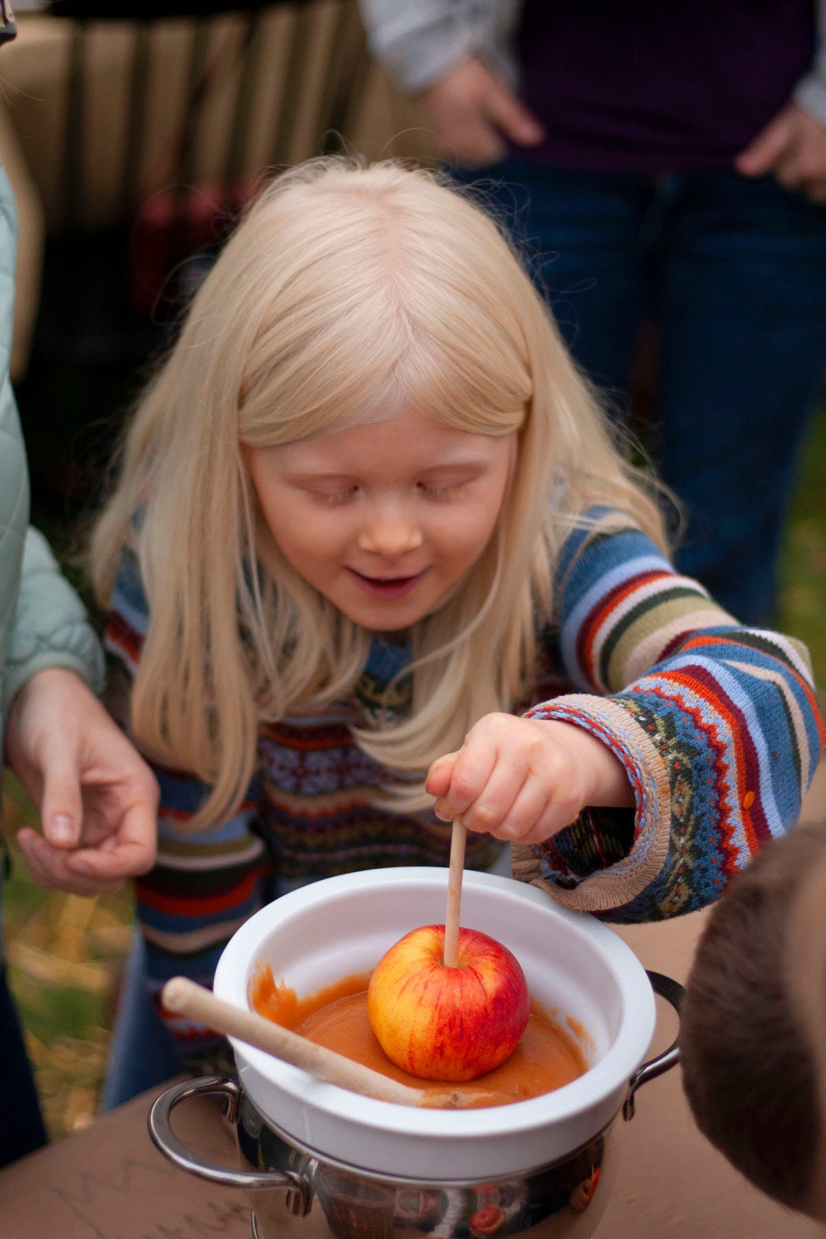 a young girl with blond hair dipping an apple in melted caramel in an outdoor setting