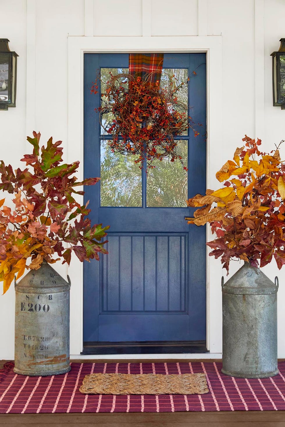 a front door decorate with a grapevine and bittersweet wreath plus buckets flanking the door that are filled with fall foliage branches