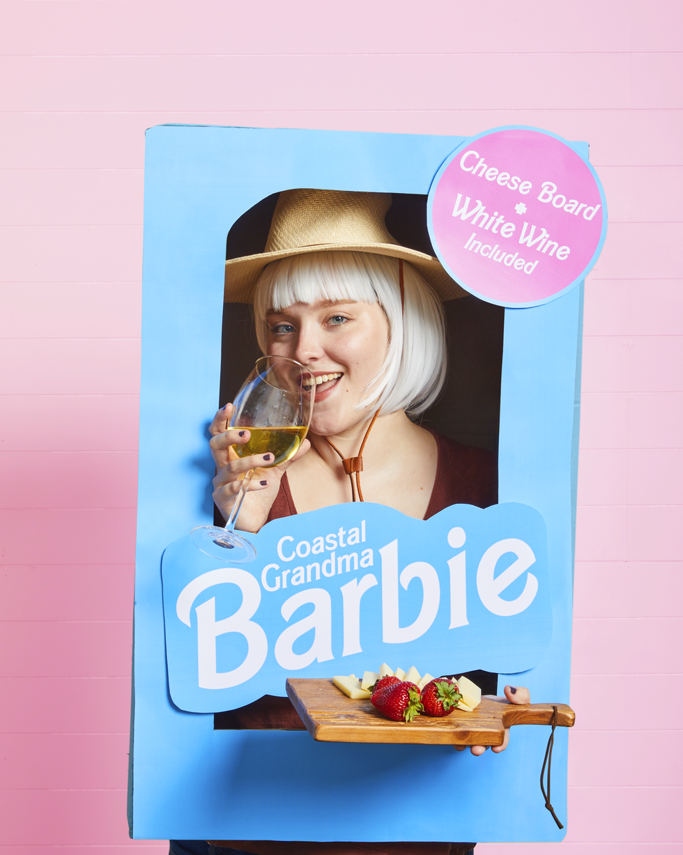 coastal grandma barbie costume featuring a woman in a blue cardboard barbie box holding a cheese plate and a glass of white wine