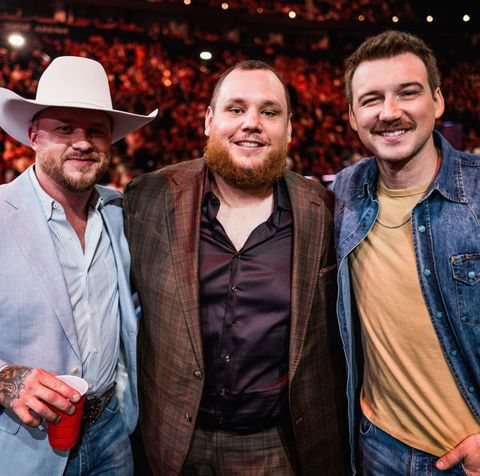 cody johnson, luke combs, and morgan wallen stand together and smile for a photo, behind them is a large audience, johnson wears a white cowboy hat and blue suit jacket, combs wears a brown plaid suit jacket, and wallen wears a jean jacket