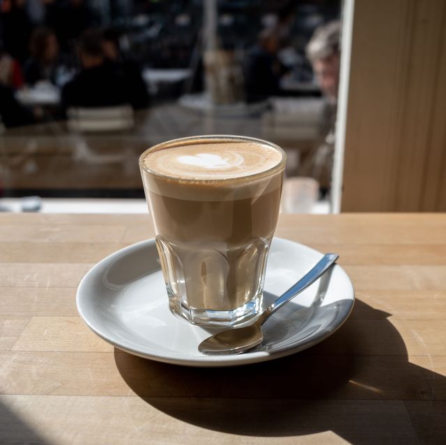 flat white coffee in clear transparent glass and white plate lay on rough wooden table counter beside windows of coffee shop and blur background of outdoor seats and people outside coffee shop