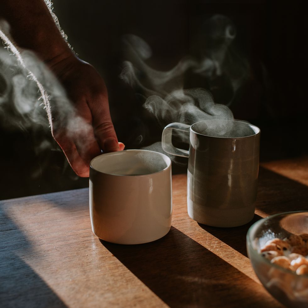 a mug of black coffee casts a shadow on a wooden dining table steam rises from the cup another cup is lifted off the table a clear bowl of breakfast cereal and milk are in frame, suggesting a morning scene of breakfast