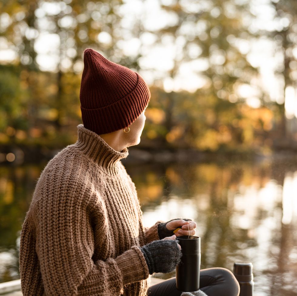 a woman preparing and drinking some fresh hot tea in a travel mug she is sitting on a small jetty on the lake in autumn
