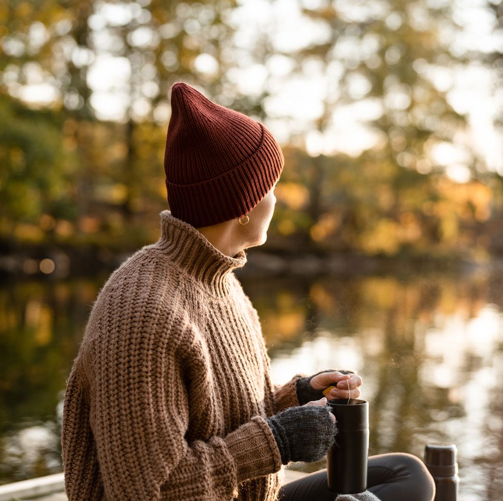 a woman preparing and drinking some fresh hot tea in a travel mug she is sitting on a small jetty on the lake in autumn