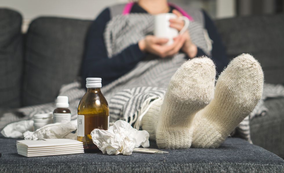 Cold medicine and sick woman drinking hot beverage to get well from flu, fever and virus. Dirty paper towels and tissues on table. Ill person wearing warm woolen stocking socks in winter.