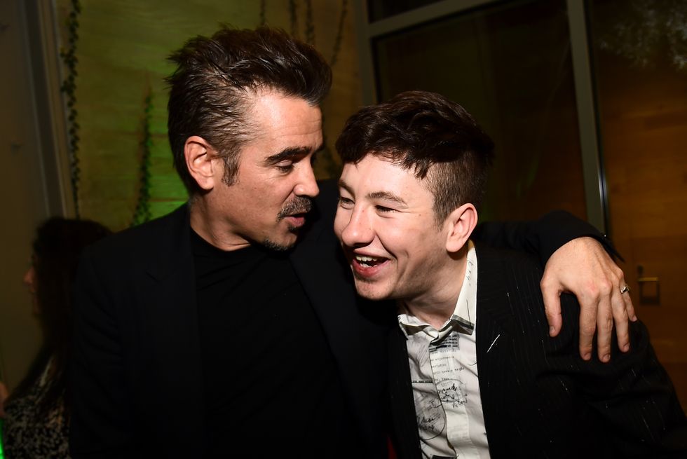colin farrell and barry keoghan stand next to own another in a dark room, farrell looks at and talks to keoghan with one arm around his shoulder, keoghan smiles