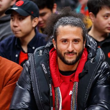 colin kaepernick smiles at the camera while sitting in a group of people, he wears a red sweatshirt and a black puffy coat, his hair is pulled back into a ponytail