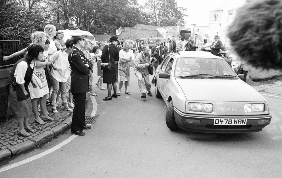 a crowd of people shouting and holding cameras follows a car