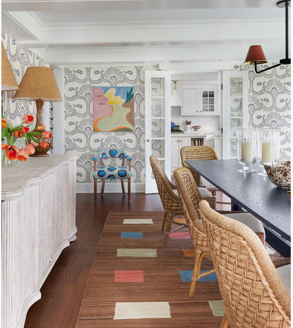 the scene features a dining table surrounded by woven chairs placed on a patterned rug that includes multicolored blocks a sideboard with a textured surface holds a vibrant bouquet of tulips one patterned chair with a blue and white motif is visible against a backdrop of intricate wallpaper a piece of abstract art hangs on the wall enhancing the rooms aesthetic in the background, there is an open doorway leading to a kitchen area with white cabinetry