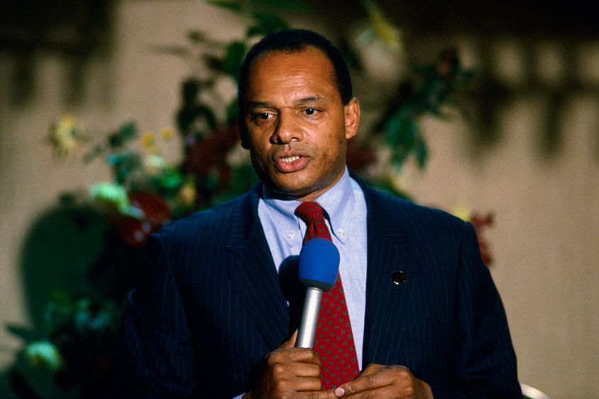 george raveling wearing a blue suit and red tie, speaking into a microphone