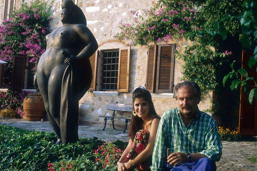 sophia vari and fernando botero sit on the ground and pose for a photo in a backyard next to a large bronze sculpture