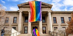 a person draped in a lgbtq flag stands in front of city hall in colorado springs, which is hanging a larger lgbtq flag