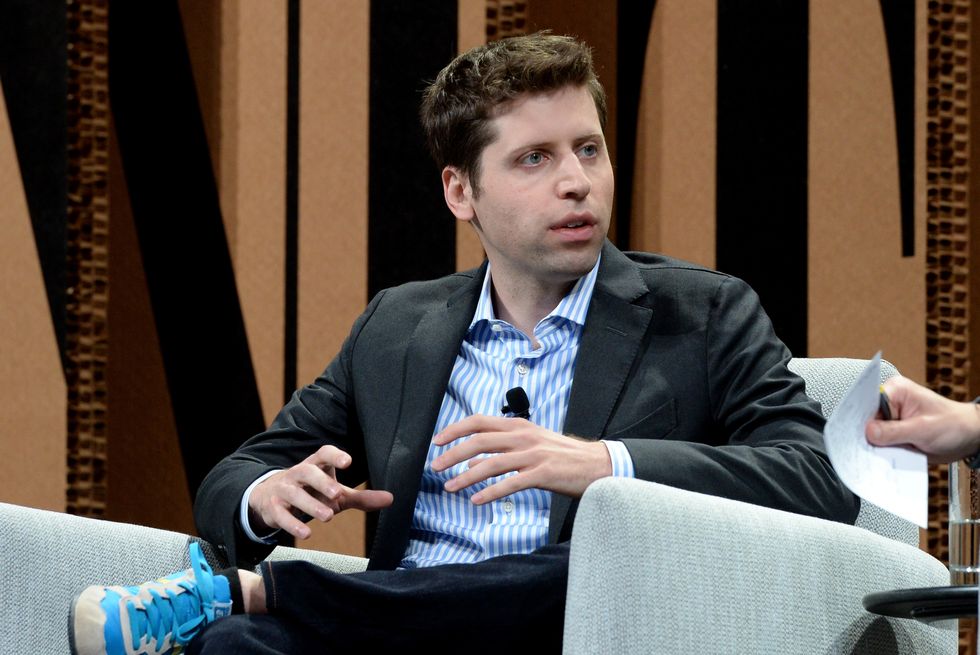 a man sits in an armchair with one leg crossed over the other, he speaks as he looks right and gestures with his hands, he wears a dark suit jacket, white and blue collared shirt, dark jeans, and blue sneakers
