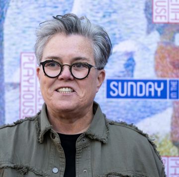 rosie o'donnell smiles and looks to the left of the camera, she wears black framed glasses, an olive green jacket and a black top, behind her is a poster of a painting