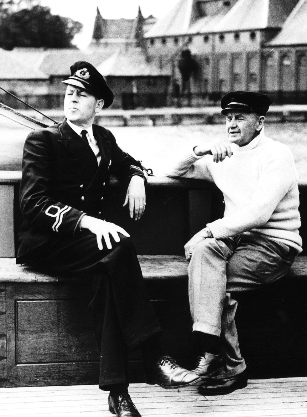 a black and white photo of charles lightoller, sitting on a bench and leaning his right arm on a railing