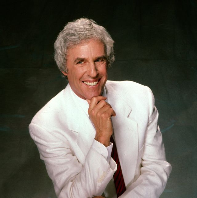 burt bacharach portrait shows the musician sitting on a stool in an all white suit with a red and blue striped tie, his right elbow is leaning on his leg and his right hand is resting underneath his chin, he smiles and looks to the right