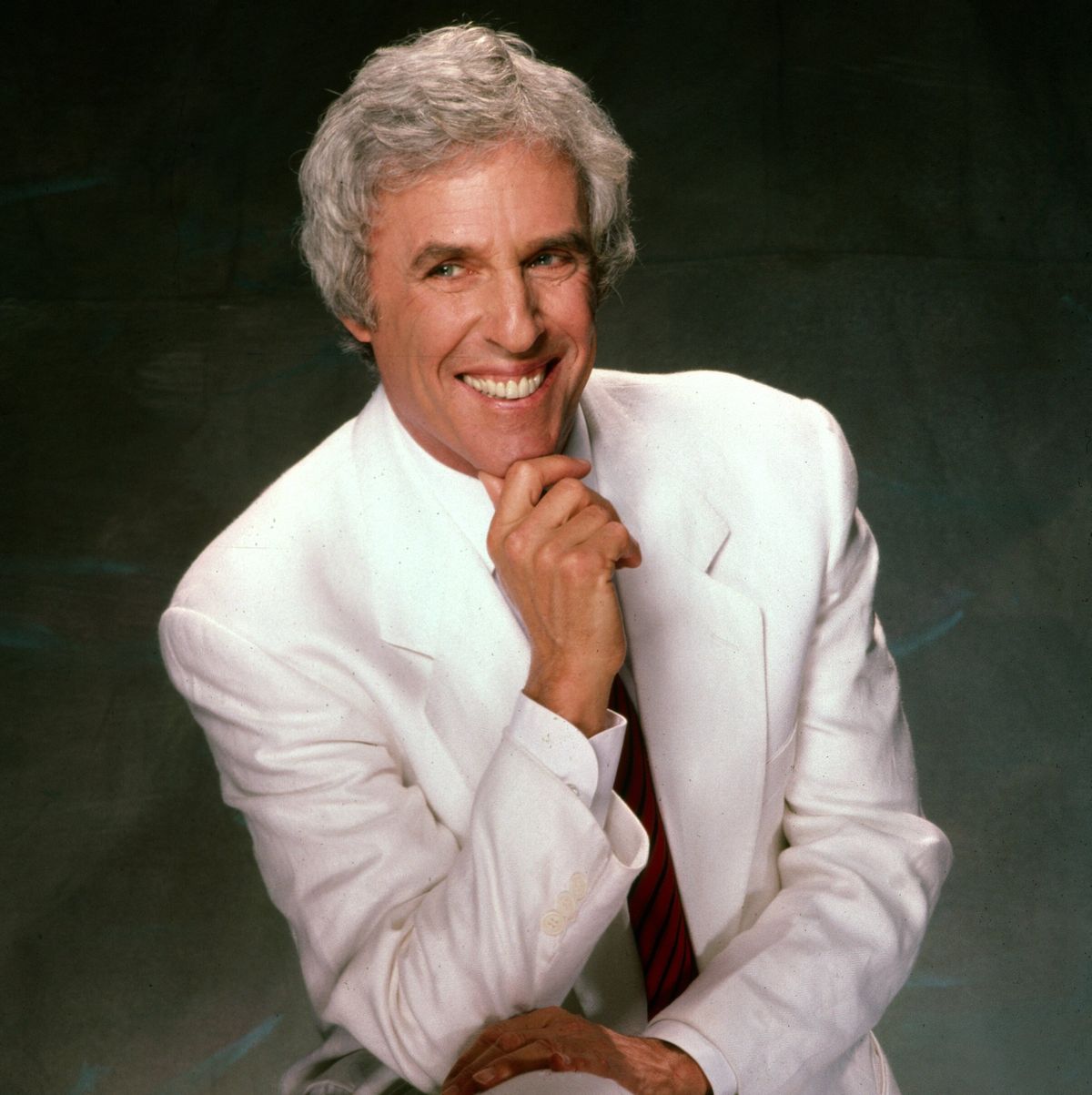 burt bacharach portrait shows the musician sitting on a stool in an all white suit with a red and blue striped tie, his right elbow is leaning on his leg and his right hand is resting underneath his chin, he smiles and looks to the right