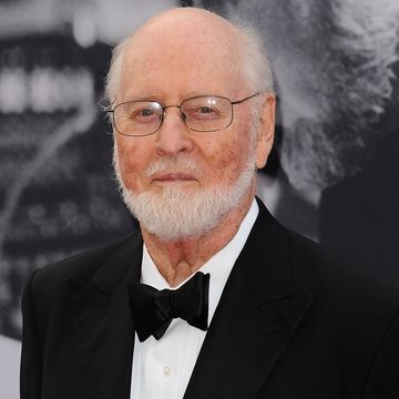 john williams smiles at the camera, he wears a black tuxedo with a white collared shirt and metal rimmed glasses