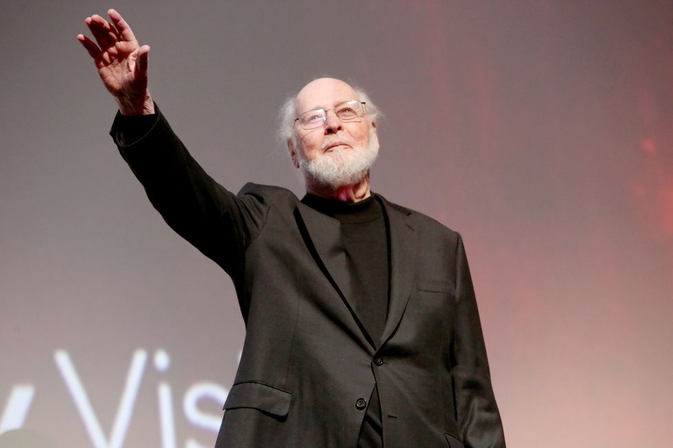 john williams, wearing a black suit jacket and shirt, waving from a stage