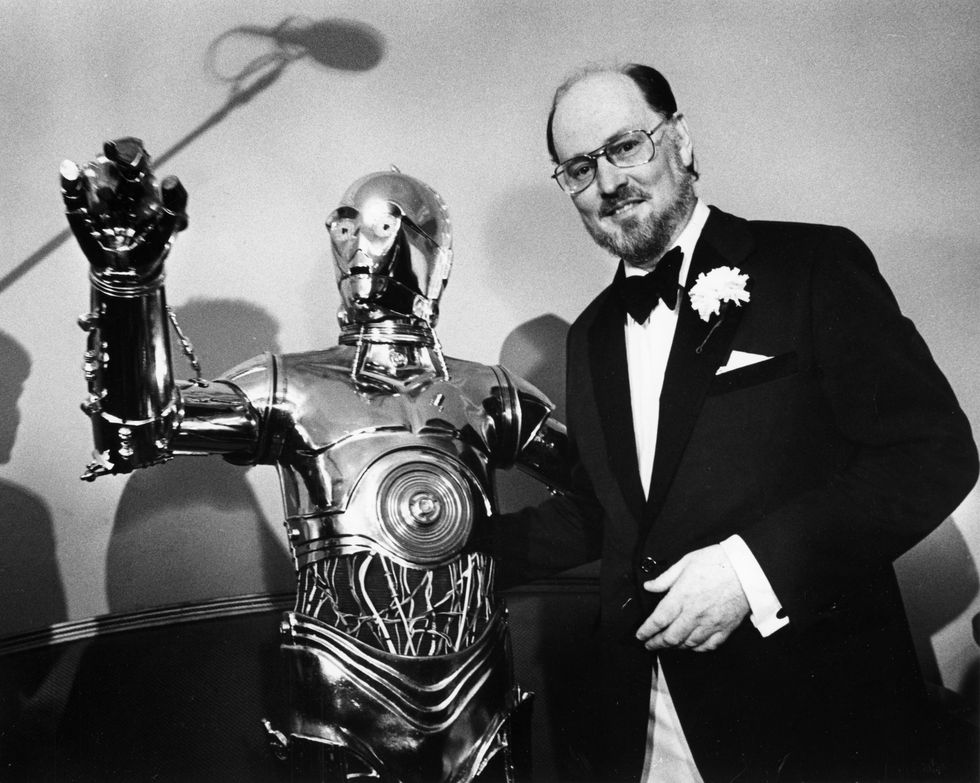 john williams smiling as he stands next to android c3po who is waving to a crowd off screen
