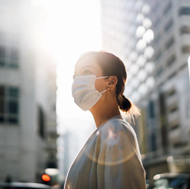 confidence and determined young asian woman with protective face mask standing in the city street, looking away with hope against beautiful lens flare in the fresh morning