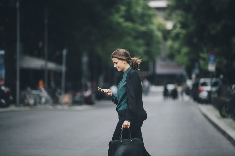 confident businesswoman using smart phone while crossing street in city