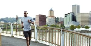 confident male athlete running on footbridge over river against clear sky in city