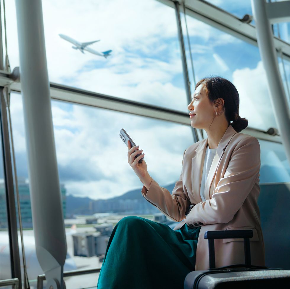 confident young asian businesswoman with suitcase looking ahead while using smartphone waiting for her flight at airport terminal. business traveller. woman in business. business lifestyle and technology. travel and vacation concept