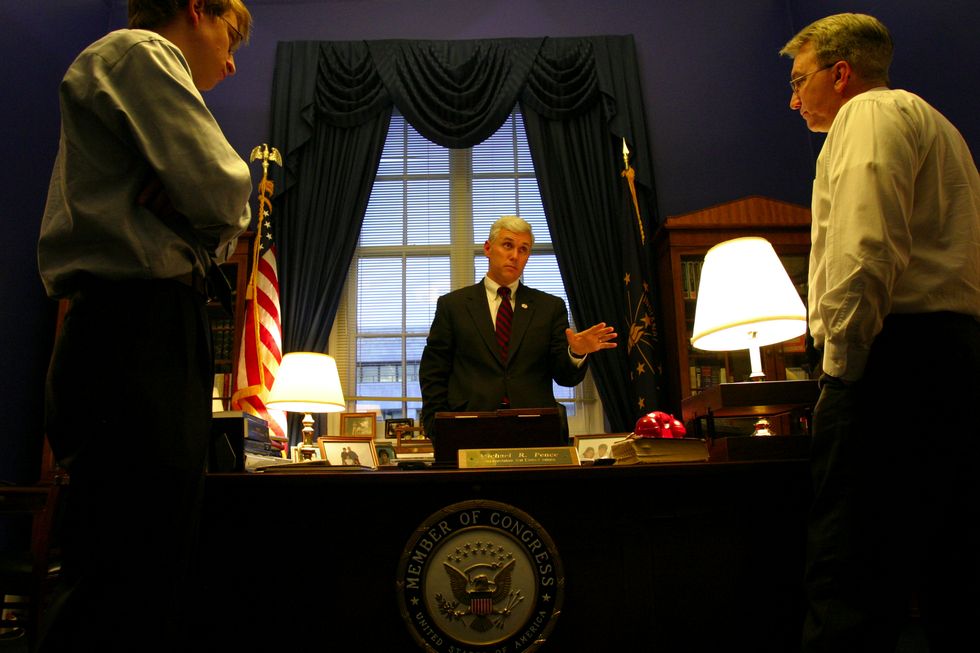 mike pence stands behind a desk with a congressional seal on the front, he looks at a man on the right of the desk as a man on the left side watches on, pence wears a suit with a white shirt and red tie, behind him is a window with ornate curtains and two flags