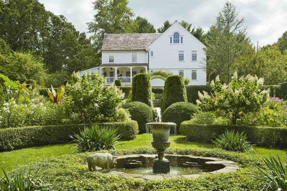 a low privet hedge forms a traditional rondel inside it, a fountain with stone from a mini quarry on the property surrounded by vinca minor ground cover a grand white farmhouse is in the background