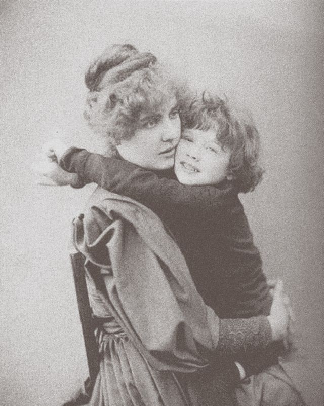 a woman in a long sleeve dress and a young boy hug, she looks to the right, he looks at the camera smiling