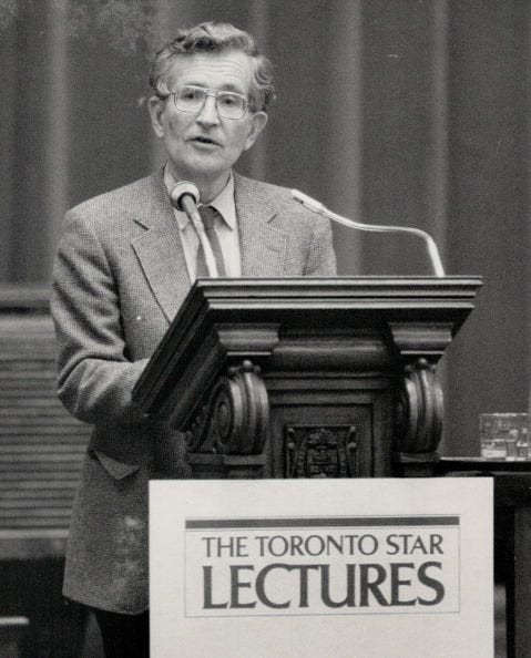 noam chomsky speaks at a wooden podium outfitted with two microphones, he wears a suit jacket, tie, dress dress, and glasses