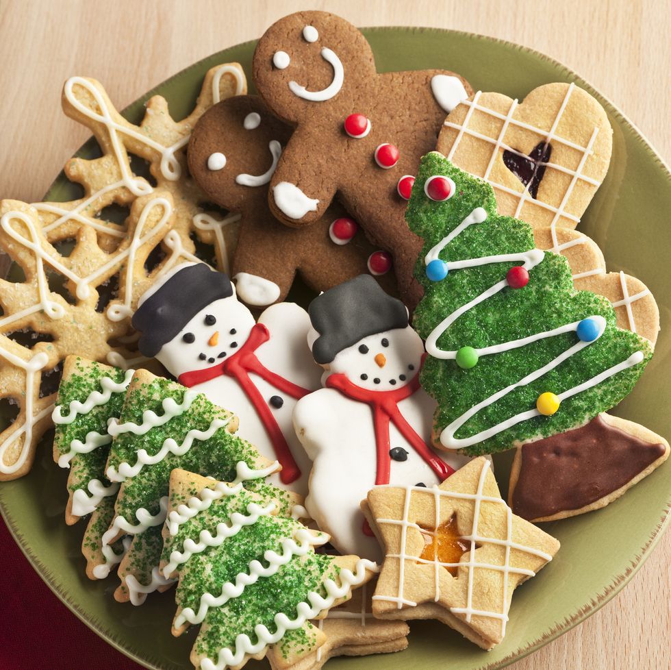 a plate filled with christmas cookies decorated with frosting and icing for winter holiday celebrations the baked dessert collection of sweet food features christmas tree, gingerbread man, snowman, star, heart, and snowflake shapes against the green background of a ceramic plate the light wooden table area allows for copy space