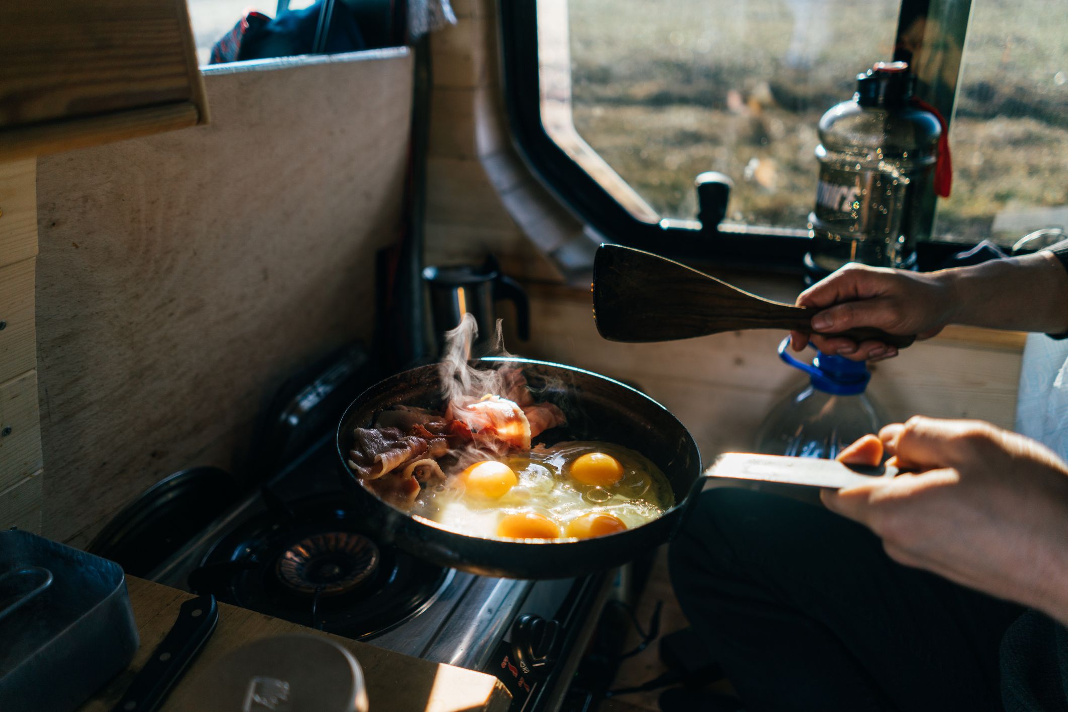 https://hips.hearstapps.com/hmg-prod/images/cooking-eggs-and-bacon-in-skillet-at-campsite-royalty-free-image-1700081441.jpg