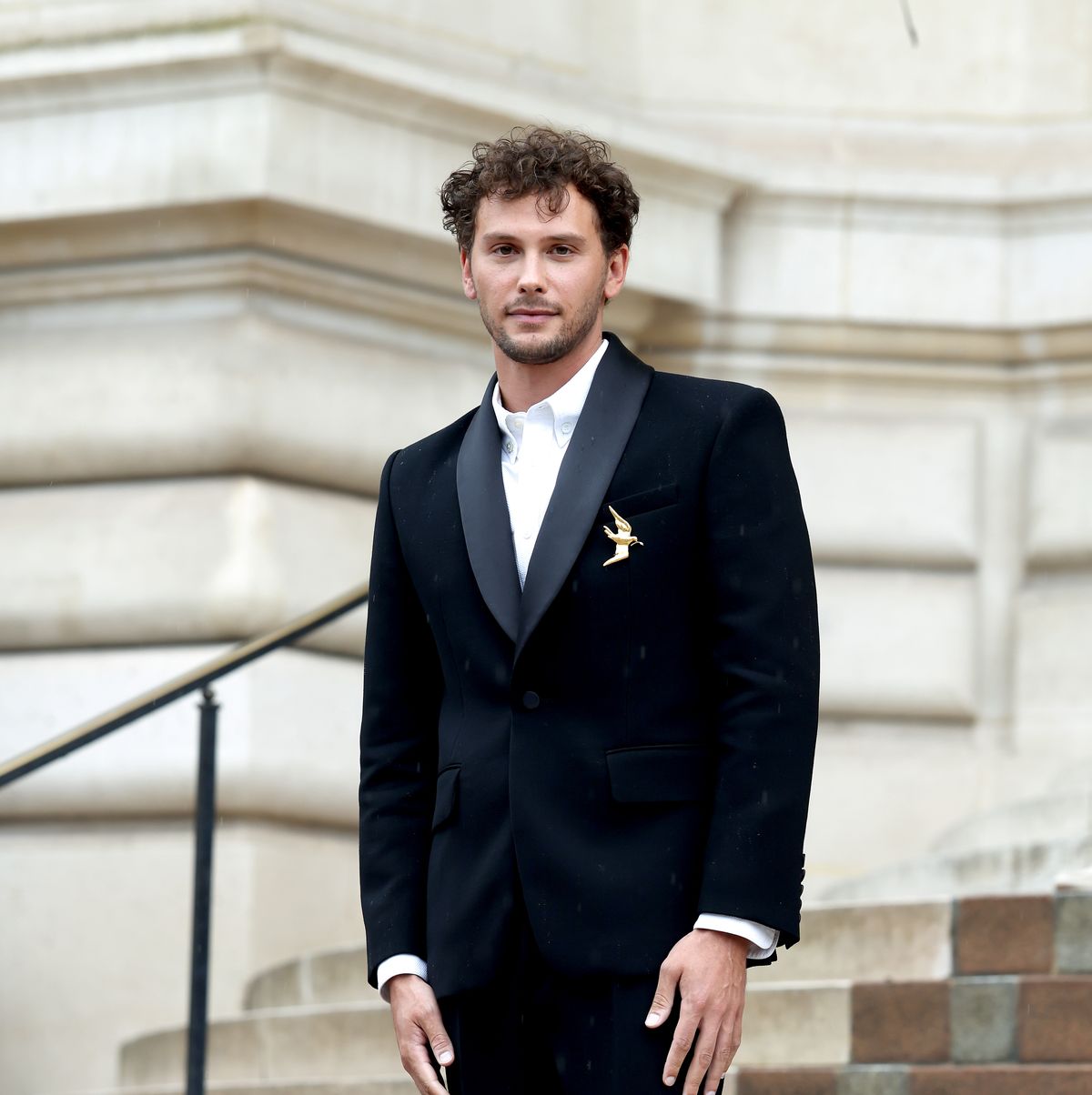 a man in a black suit with a golden bird pin stands on concrete steps outside a building and looks at the camera