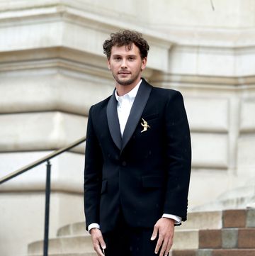 a man in a black suit with a golden bird pin stands on concrete steps outside a building and looks at the camera