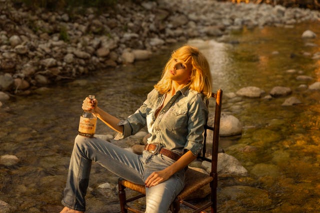 a person seated on a wooden chair by a river holding a bottle