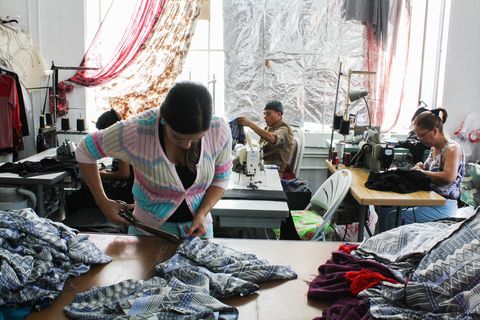 a woman cuts fabric at a warehouse, behind her other workers labor at sewing machines