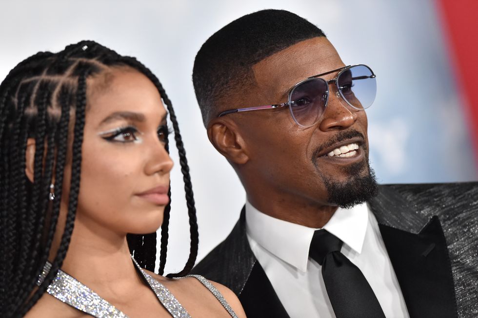 corinne foxx, wearing a gray dress, and jamie foxx, wearing a black and grey suit and tie and sunglasses, look off camera in the same direction