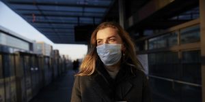 Young woman standing on train station wearing protective mask, using phone