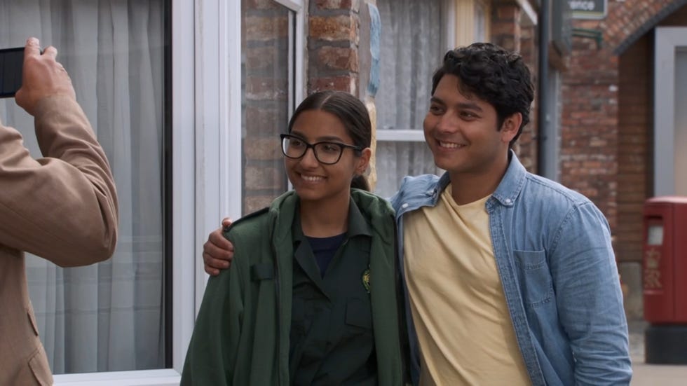coronation street asha and aadi alahan in the street having a photo taken by dev, asha is in her green parademic uniform while aadi is in a yellow tshirt with a blue thin open shirt, and has his arm around her