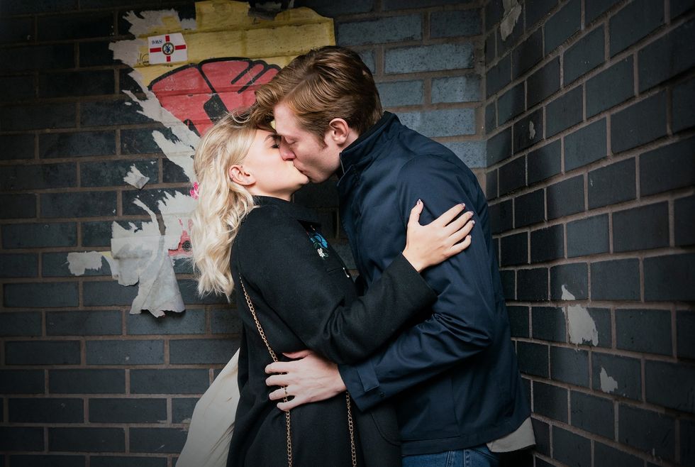 in a scene from coronation street, bethany and daniel kiss in front of a grey brick wall with a partially removed, brightly coloured poster on it