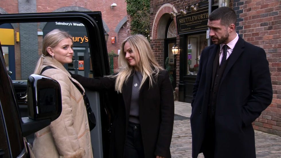 three individuals engaged in conversation near a vehicle in an urban setting with brick buildings