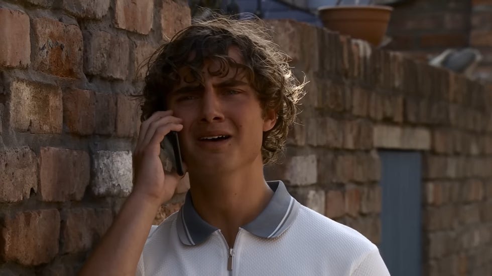 coronation street, brody in a white tshirt leaning against a brick wall looking devastated while on the phone