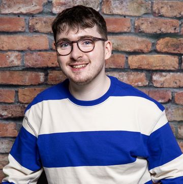 jack carroll as bobby in coronation street, a young man stands with a walking aid and smiles, he wears glasses and a blue and white striped top