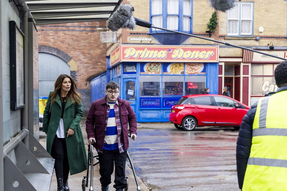 Bobby y Carla en Coronation Street, un detrás de escena de la filmación mientras una mujer y un joven con ayuda para caminar caminan por la calle.