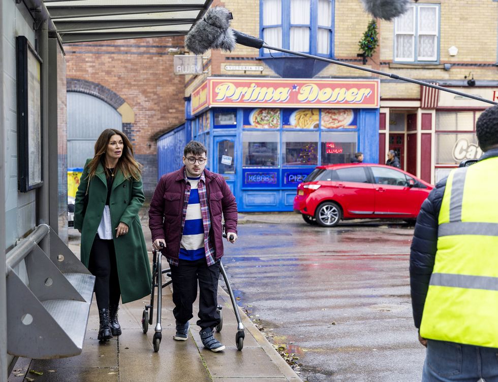 Bobby und Carla in der Coronation Street, ein Blick hinter die Kulissen der Dreharbeiten, während eine Frau und ein junger Mann mit Gehhilfe die Straße entlang schlendern