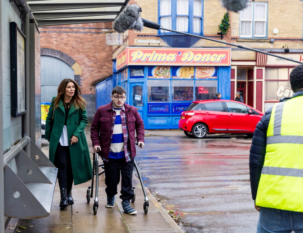 Bobby und Carla in der Coronation Street, ein Blick hinter die Kulissen der Dreharbeiten, während eine Frau und ein junger Mann mit Gehhilfe die Straße entlang schlendern