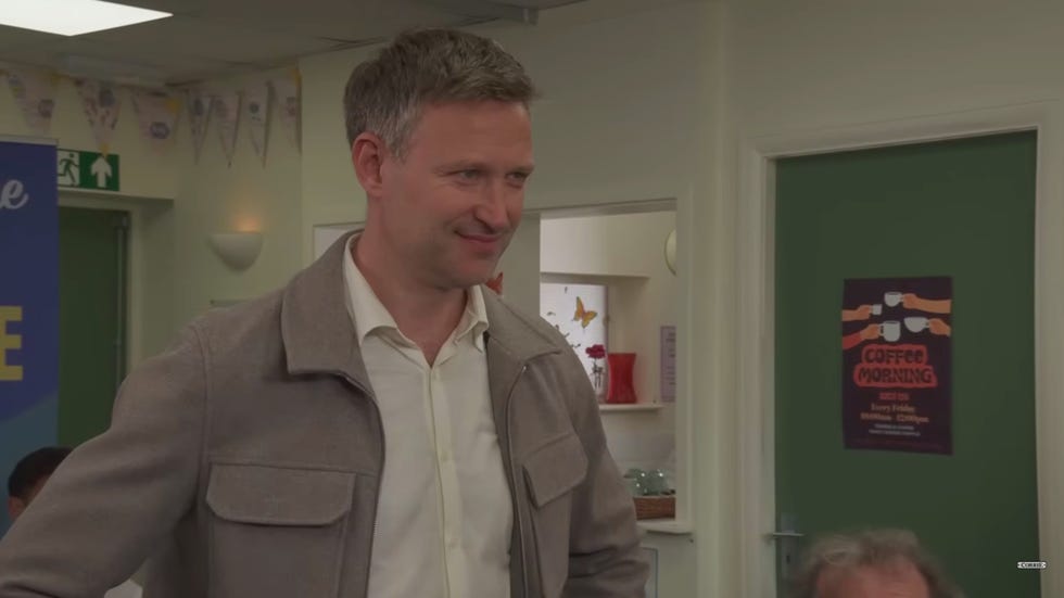 coronation street, noah with an evil smile wearing a white shirt and grey jacket inside the community centre
